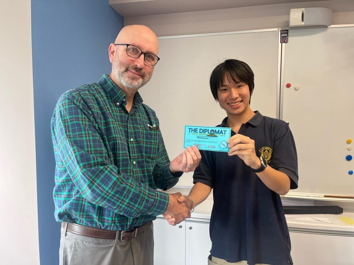 Student editor Rinnosuke('27) receives his 50-published-stories certificate and pin from Mr. Douglas